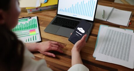Businesswoman synchronizes mobile phone and laptop data using new program. Lady sitting at table with documents and statistics transfers data to cloud for information security