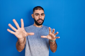 Poster - Middle east man with beard standing over blue background afraid and terrified with fear expression stop gesture with hands, shouting in shock. panic concept.