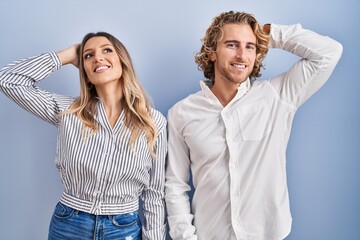 Wall Mural - Young couple standing over blue background smiling confident touching hair with hand up gesture, posing attractive and fashionable