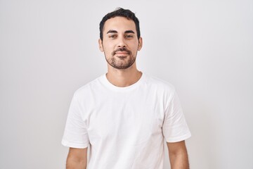 Wall Mural - Handsome hispanic man standing over white background relaxed with serious expression on face. simple and natural looking at the camera.