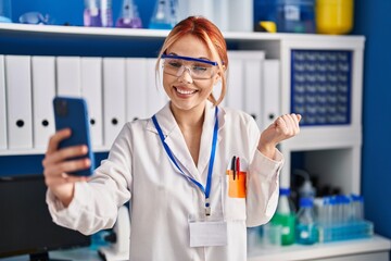 Canvas Print - Young caucasian woman working at scientist laboratory doing video call with smartphone screaming proud, celebrating victory and success very excited with raised arm