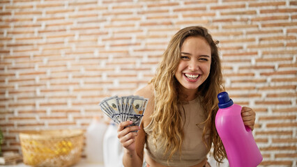 Poster - Young beautiful hispanic woman holding detergent bottle and dollars at laundry room