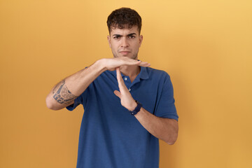 Sticker - Young hispanic man standing over yellow background doing time out gesture with hands, frustrated and serious face