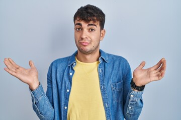 Sticker - Young hispanic man standing over blue background clueless and confused expression with arms and hands raised. doubt concept.