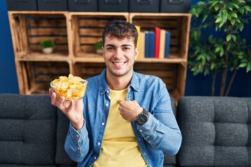 Sticker - Young hispanic man holding potato chips pointing finger to one self smiling happy and proud