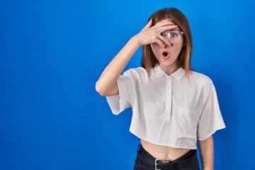 Wall Mural - Beautiful woman standing over blue background peeking in shock covering face and eyes with hand, looking through fingers with embarrassed expression.