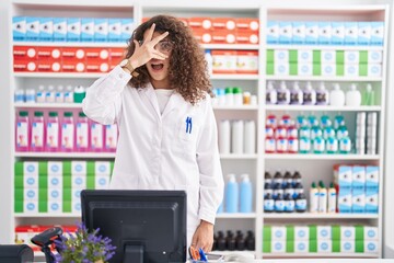 Sticker - Hispanic woman with curly hair working at pharmacy drugstore peeking in shock covering face and eyes with hand, looking through fingers with embarrassed expression.