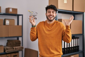 Poster - Hispanic man with beard standing by manikin at small business holding cart celebrating victory with happy smile and winner expression with raised hands