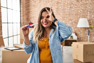 Wall Mural - Young hispanic woman holding pregnancy test result at new home smiling happy doing ok sign with hand on eye looking through fingers