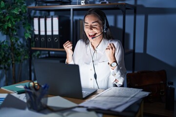 Poster - Young brunette woman wearing call center agent headset working late at night very happy and excited doing winner gesture with arms raised, smiling and screaming for success. celebration concept.