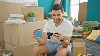 Poster - Young hispanic man watching video on smartphone drinking coffee at new home