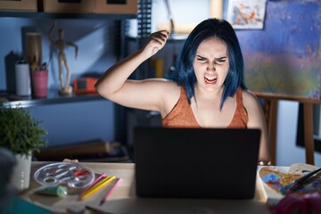 Poster - Young modern girl with blue hair sitting at art studio with laptop at night angry and mad raising fist frustrated and furious while shouting with anger. rage and aggressive concept.