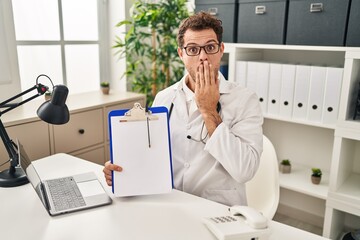 Poster - Young hispanic man wearing doctor stethoscope holding clipboard covering mouth with hand, shocked and afraid for mistake. surprised expression