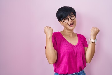 Poster - Young asian woman with short hair standing over pink background celebrating surprised and amazed for success with arms raised and eyes closed. winner concept.