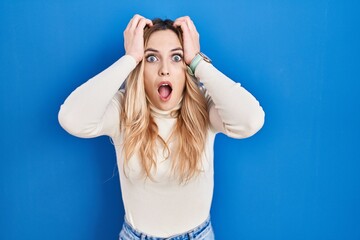 Wall Mural - Young caucasian woman standing over blue background crazy and scared with hands on head, afraid and surprised of shock with open mouth