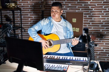 Canvas Print - Hispanic young man playing classic guitar at music studio puffing cheeks with funny face. mouth inflated with air, crazy expression.