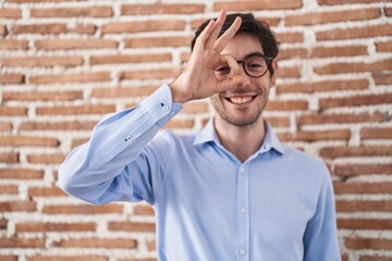 Poster - Young hispanic man standing over brick wall background doing ok gesture with hand smiling, eye looking through fingers with happy face.