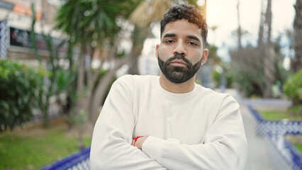 Sticker - Young hispanic man standing with serious expression and arms crossed gesture at park