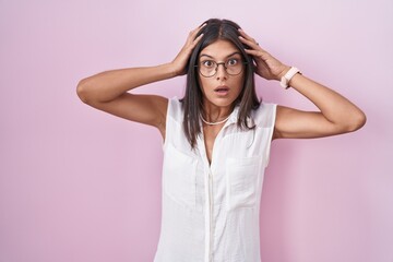 Poster - Brunette young woman standing over pink background wearing glasses crazy and scared with hands on head, afraid and surprised of shock with open mouth