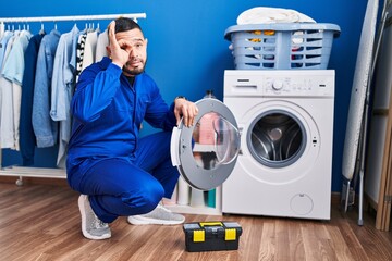 Wall Mural - Hispanic repairman working on washing machine smiling happy doing ok sign with hand on eye looking through fingers