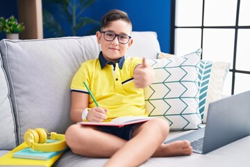 Sticker - Young hispanic kid doing homework sitting on the sofa approving doing positive gesture with hand, thumbs up smiling and happy for success. winner gesture.