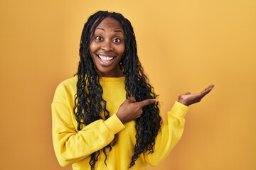 Sticker - African woman standing over yellow background amazed and smiling to the camera while presenting with hand and pointing with finger.