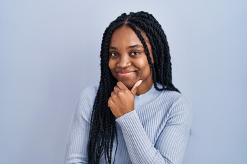 Wall Mural - African american woman standing over blue background looking confident at the camera smiling with crossed arms and hand raised on chin. thinking positive.