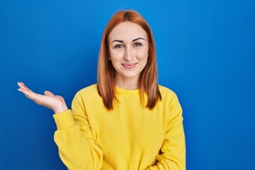 Wall Mural - Young woman standing over blue background smiling cheerful presenting and pointing with palm of hand looking at the camera.