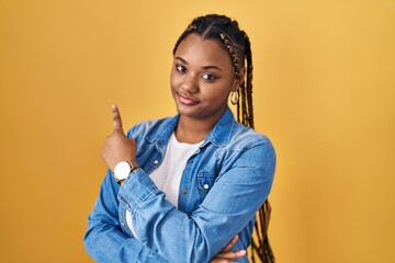 Sticker - African american woman with braids standing over yellow background pointing with hand finger to the side showing advertisement, serious and calm face