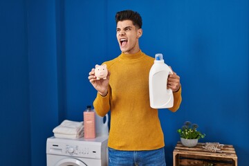 Poster - Young hispanic man holding detergent bottle and piggy bank angry and mad screaming frustrated and furious, shouting with anger. rage and aggressive concept.