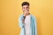 Young hispanic man with tattoos standing over yellow background smelling something stinky and disgusting, intolerable smell, holding breath with fingers on nose. bad smell