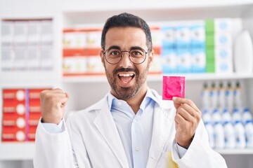 Sticker - Hispanic man with beard working at pharmacy drugstore holding condom screaming proud, celebrating victory and success very excited with raised arms