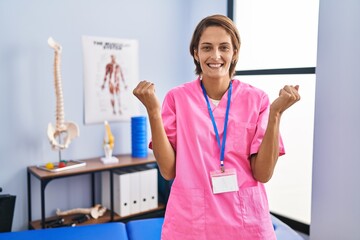 Poster - Brunette woman working at rehabilitation clinic celebrating surprised and amazed for success with arms raised and open eyes. winner concept.