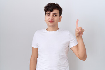 Poster - Young non binary man wearing casual white t shirt showing and pointing up with finger number one while smiling confident and happy.