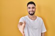 © Krakenimages.com - Young handsome man wearing casual t shirt over yellow background smiling friendly offering handshake as greeting and welcoming. successful business.