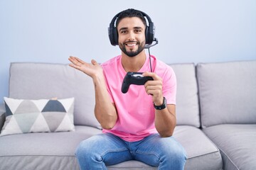 Poster - Hispanic young man playing video game holding controller sitting on the sofa pointing aside with hands open palms showing copy space, presenting advertisement smiling excited happy