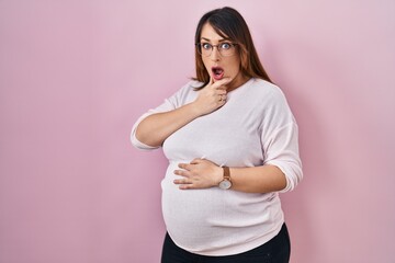 Poster - Pregnant woman standing over pink background looking fascinated with disbelief, surprise and amazed expression with hands on chin