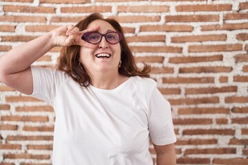 Sticker - Senior woman with glasses standing over bricks wall doing peace symbol with fingers over face, smiling cheerful showing victory