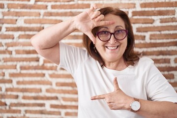 Sticker - Senior woman with glasses standing over bricks wall smiling making frame with hands and fingers with happy face. creativity and photography concept.