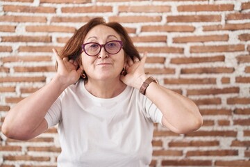 Sticker - Senior woman with glasses standing over bricks wall trying to hear both hands on ear gesture, curious for gossip. hearing problem, deaf