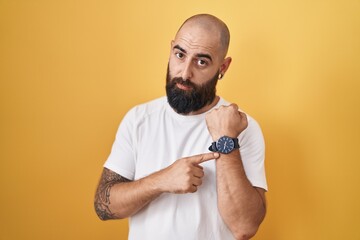 Wall Mural - Young hispanic man with beard and tattoos standing over yellow background in hurry pointing to watch time, impatience, looking at the camera with relaxed expression