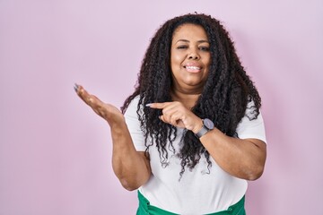 Poster - Plus size hispanic woman standing over pink background amazed and smiling to the camera while presenting with hand and pointing with finger.