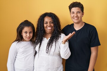 Poster - Family of mother, daughter and son standing over yellow background smiling cheerful presenting and pointing with palm of hand looking at the camera.