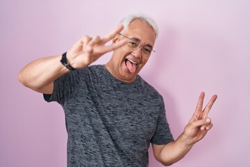 Poster - Middle age man with grey hair standing over pink background smiling with tongue out showing fingers of both hands doing victory sign. number two.