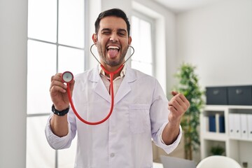 Canvas Print - Young hispanic doctor man with beard holding stethoscope auscultating sticking tongue out happy with funny expression.