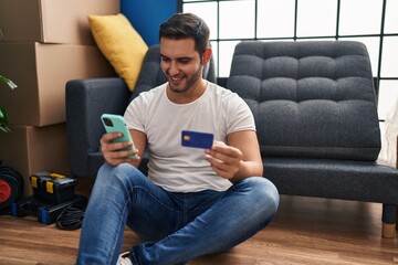 Wall Mural - Young hispanic man using smartphone and credit card sitting on floor at new home