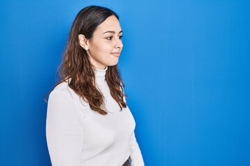 Sticker - Young hispanic woman standing over blue background looking to side, relax profile pose with natural face and confident smile.