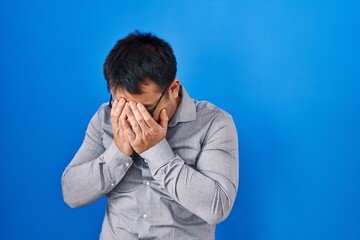 Sticker - Young chinese man standing over blue background with sad expression covering face with hands while crying. depression concept.