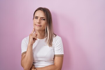 Poster - Blonde caucasian woman standing over pink background thinking concentrated about doubt with finger on chin and looking up wondering