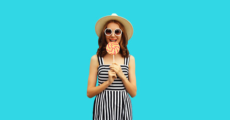 Portrait of happy young woman with colorful lollipop wearing summer straw hat, sunglasses, striped dress on blue background